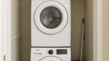 A laundry room with two stacked washing machines, a shelf with laundry supplies, a basket, and a mop.