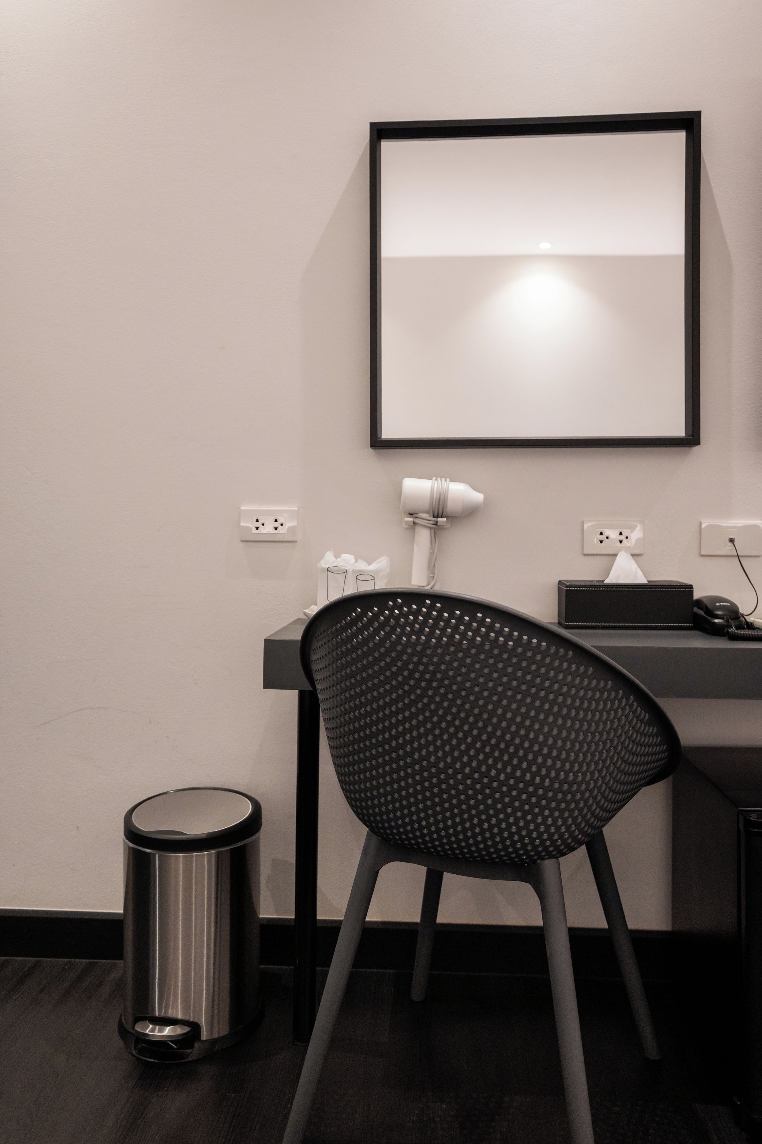 A modern office chair with a perforated backrest, a desk with a computer and a lamp, a trash can, and a framed picture on the wall.