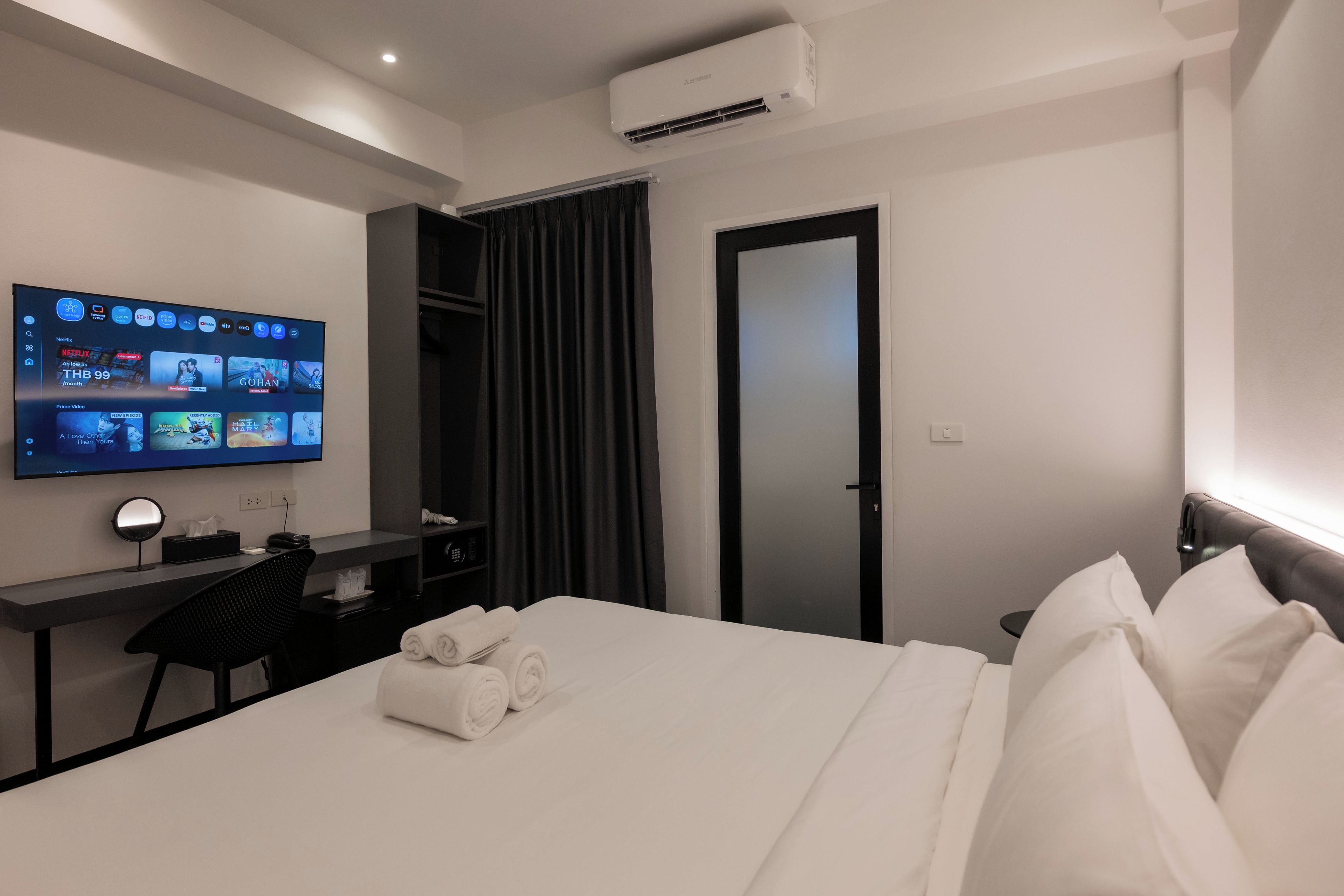 Una habitación de hotel con una cama, un escritorio con una computadora, un televisor montado en la pared y un equipo de aire acondicionado.