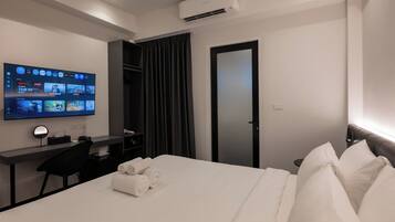 A hotel room with a bed, a desk with a computer, a TV mounted on the wall, and an air conditioning unit.