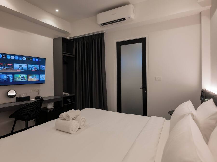 A hotel room with a bed, a desk with a computer, a TV mounted on the wall, and an air conditioning unit.