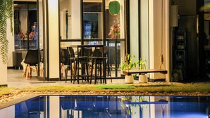 Un área interior de piscina moderna con cerramiento de vidrio, una zona de comedor con sillas y mesas, y un pequeño jardín con plantas.