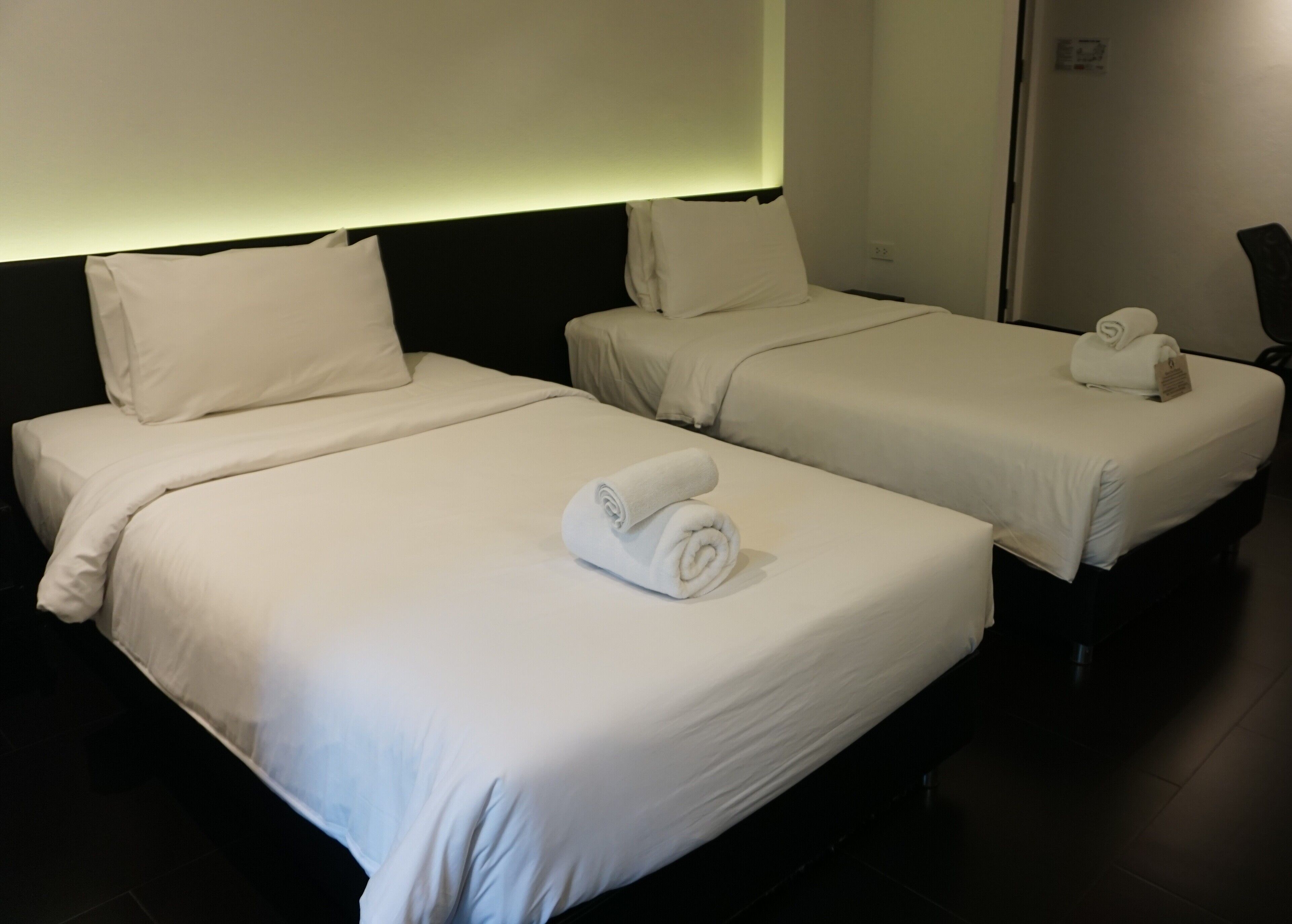 Two adjacent hotel beds with white linens and rolled towels, a dark headboard, and a wall-mounted light.