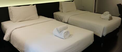 Two adjacent hotel beds with white linens and rolled towels, a dark headboard, and a wall-mounted light.
