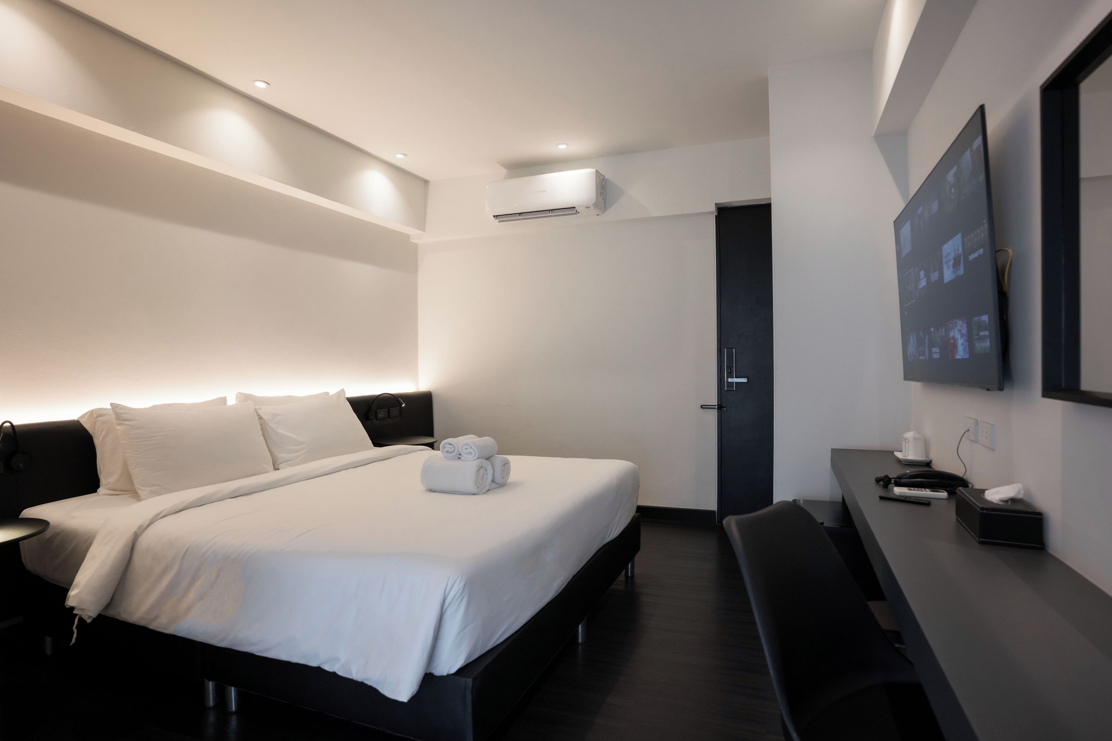 A modern hotel room with a neatly made bed, a desk with a chair, a flat-screen TV mounted on the wall, and an air conditioning unit above the bed.