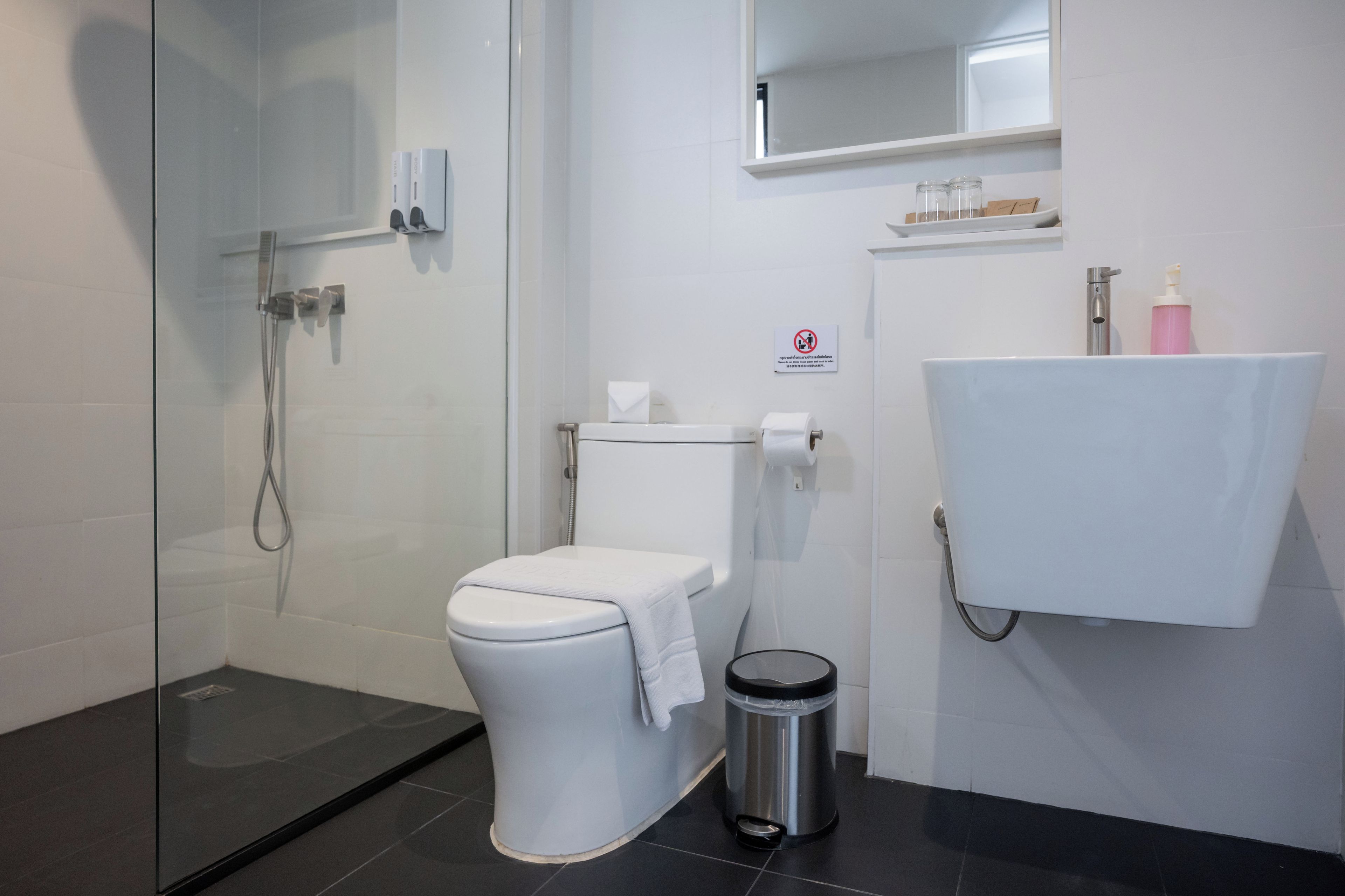 Een moderne badkamer met een douchecabine van glas, een toilet, een wastafel met zeepdispenser en een prullenbak.