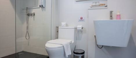 A modern bathroom with a glass shower enclosure, a toilet, a sink with a soap dispenser, and a trash bin.