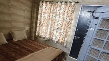 Une chambre avec un mur en bois, un lit recouvert d’une couverture marron, des rideaux à motifs floraux et une fenêtre donnant sur un immeuble.