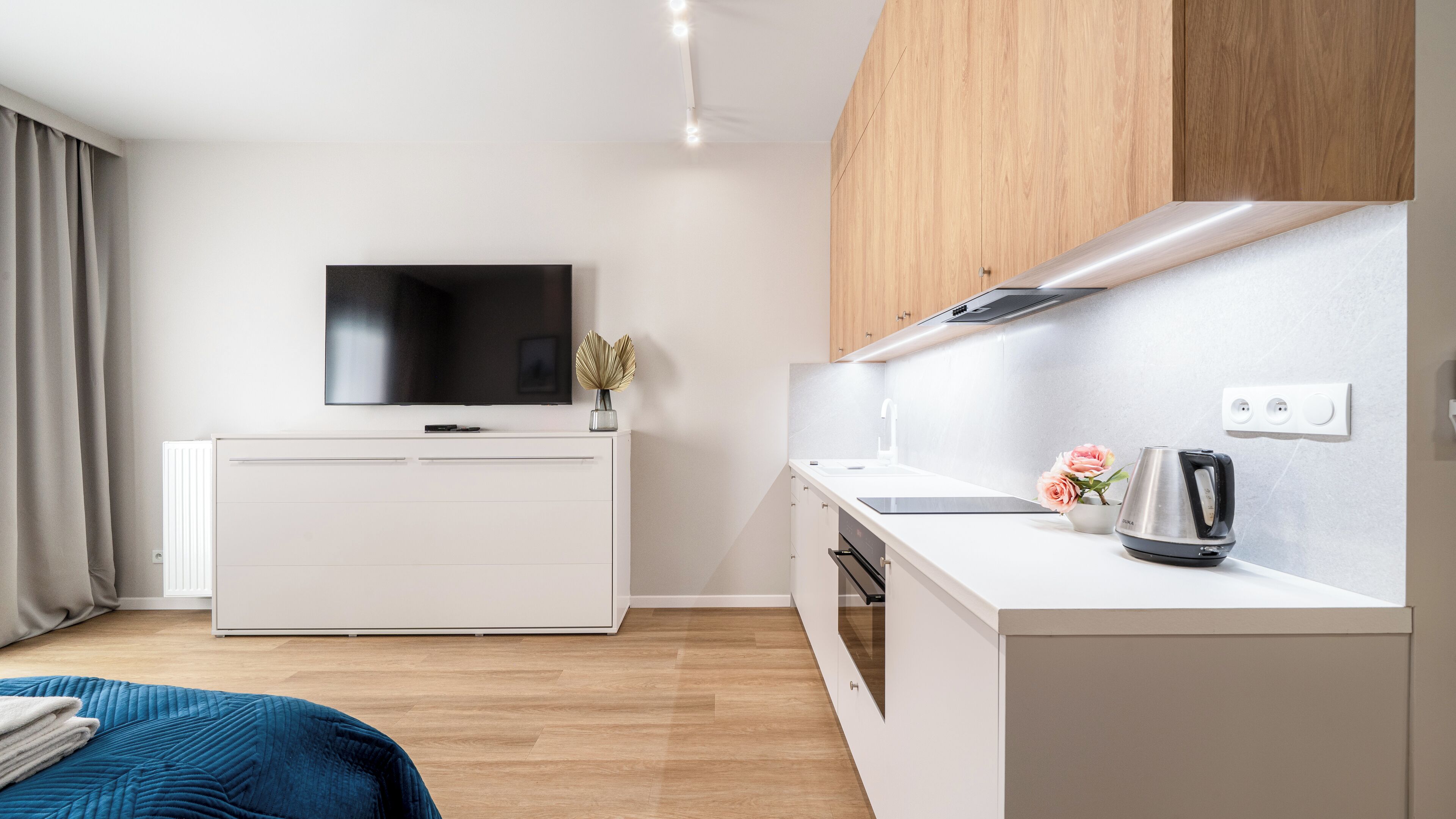 A modern hotel room with a flat-screen TV, a white cabinet, a kitchen area with a kettle, and a bed with a blue blanket.