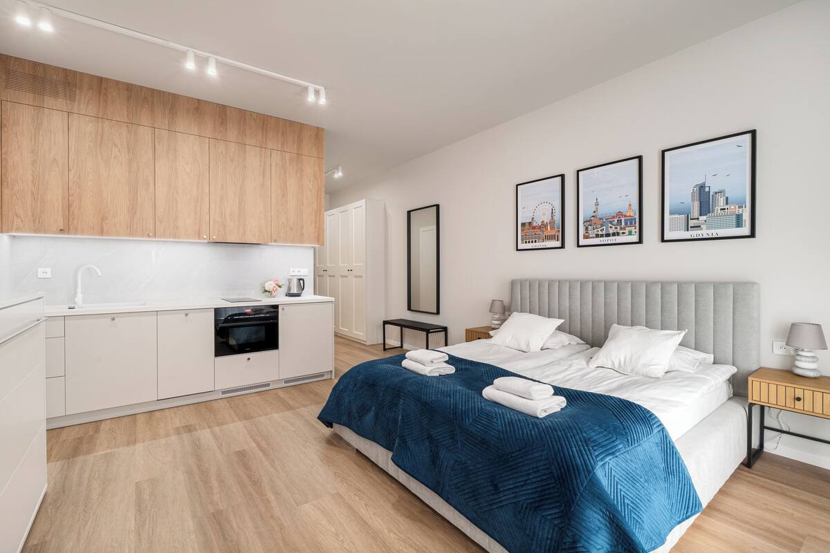 A modern hotel room with a kitchenette, a bed with a blue bedspread, a nightstand, and framed cityscape pictures on the wall.