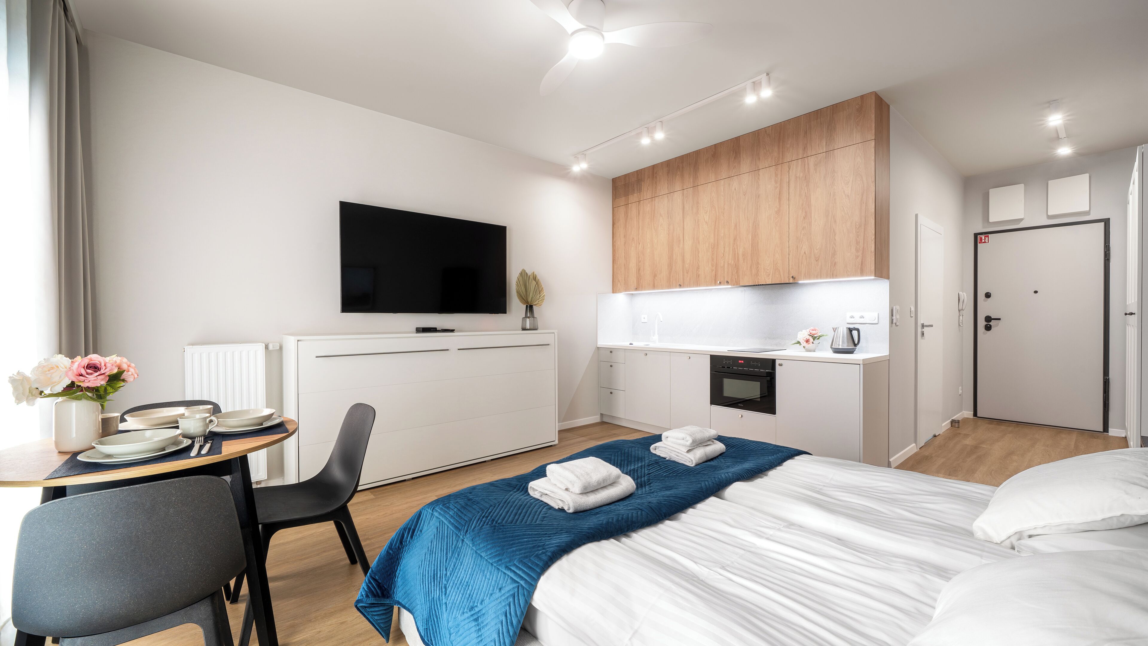 Ett modernt hotellrum med en säng, ett skrivbord, en stol, en TV, ett litet kök och en dörr.