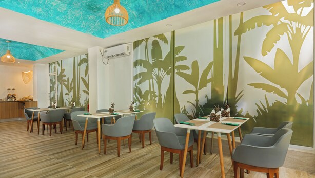 Un espace repas moderne avec un plancher en bois, des chaises et des tables grises. On y trouve des luminaires décoratifs au plafond et une fresque murale représentant un feuillage tropical.
