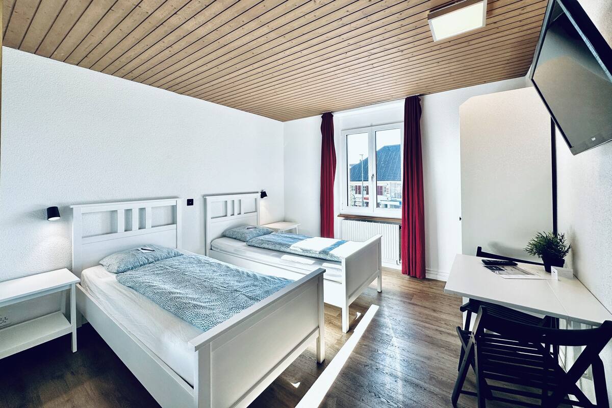 Una habitació d'hotel moderna amb dos llits individuals, un escriptori, una cadira, un televisor i una finestra amb cortines vermelles.