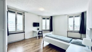 A modern, well-lit room with a wooden floor, a bed, a desk with a chair, and a TV mounted on the wall.