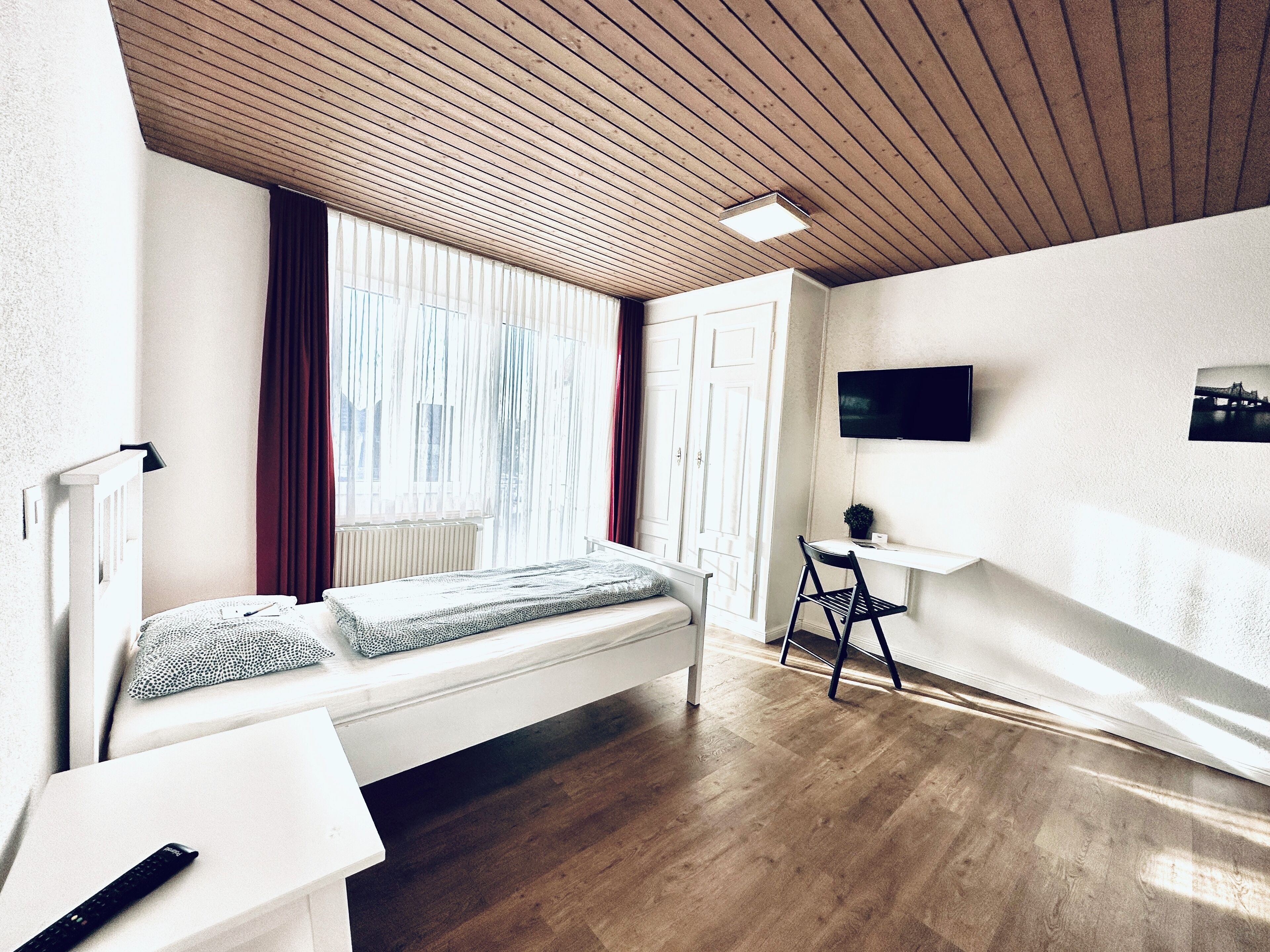 Un dormitorio con techo de madera, cama con cabecera blanca, escritorio con silla, televisor montado en la pared y ventana con cortinas.