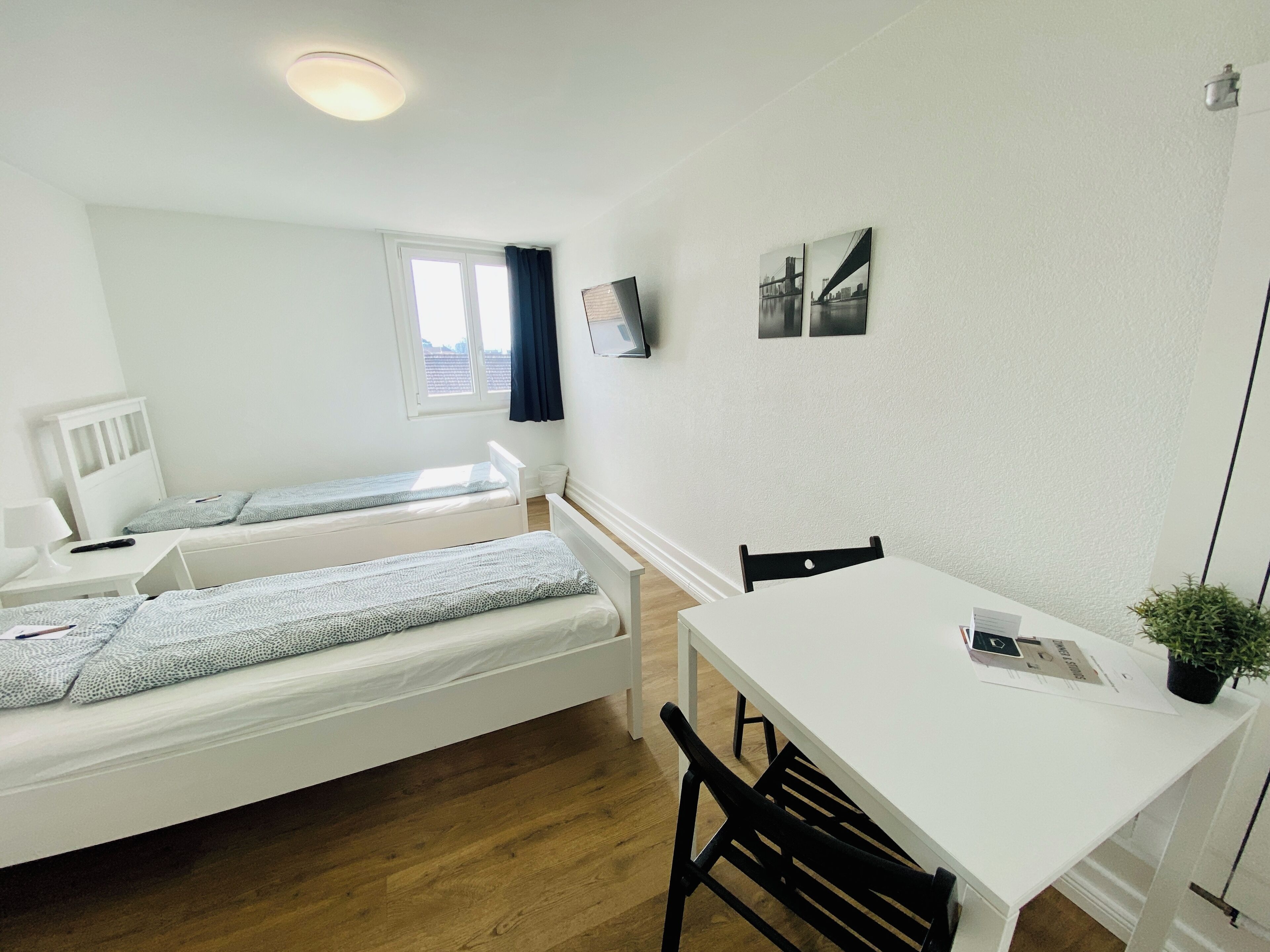 Une petite chambre propre, avec deux lits simples, un bureau blanc, une chaise, un téléviseur fixé au mur et une fenêtre avec des rideaux.