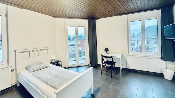 A bedroom with a bed, a desk, and a chair. There is a window with a view of buildings.