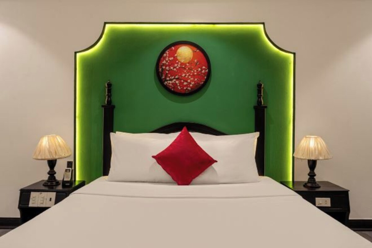 A hotel room with a green headboard, a white bed with a red pillow, and two bedside tables with lamps.