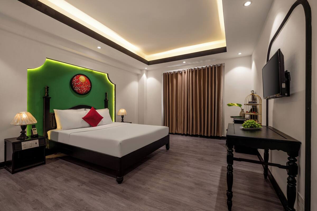 Una habitación de hotel moderna con una cama grande, una pared de acento verde, un televisor de pantalla plana y una pequeña mesa con un florero y fruta.