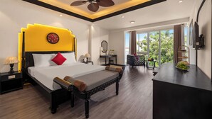 A modern hotel room with a large bed, a ceiling fan, a desk with a fruit bowl, and a view of greenery outside. - Fusion Point Hotel (Hue)