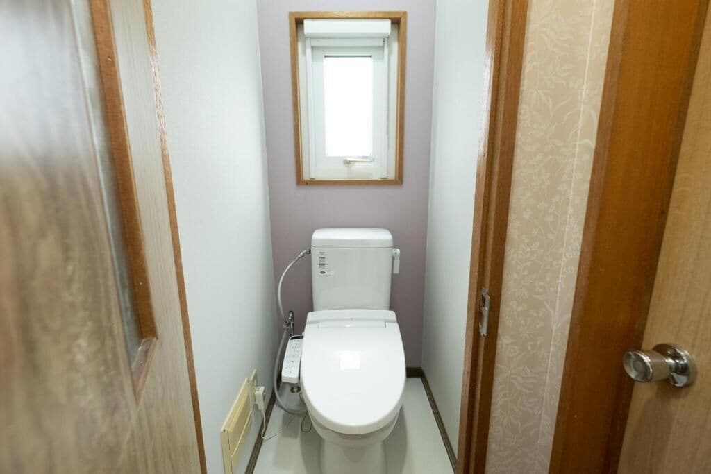 Un baño pequeño y estrecho con inodoro, una ventana en lo alto y la puerta a la derecha.