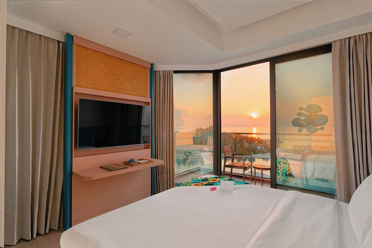 A hotel room with a large bed, a TV mounted on a wall, a balcony with a view of the sunset, and a small table with a chair.