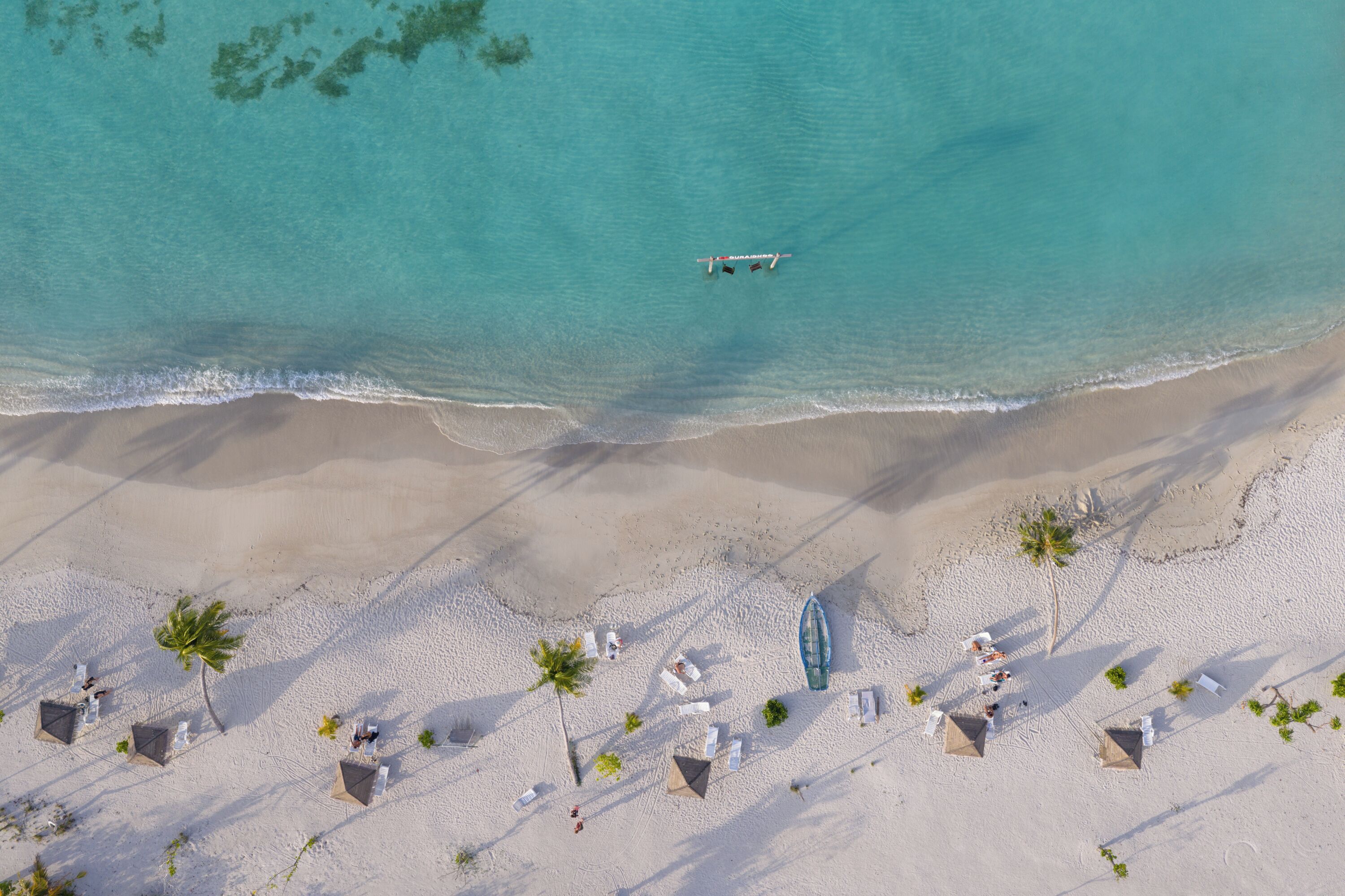 An aerial view of a beach with clear turquoise water, a boat, and a beach area with umbrellas and lounge chairs.