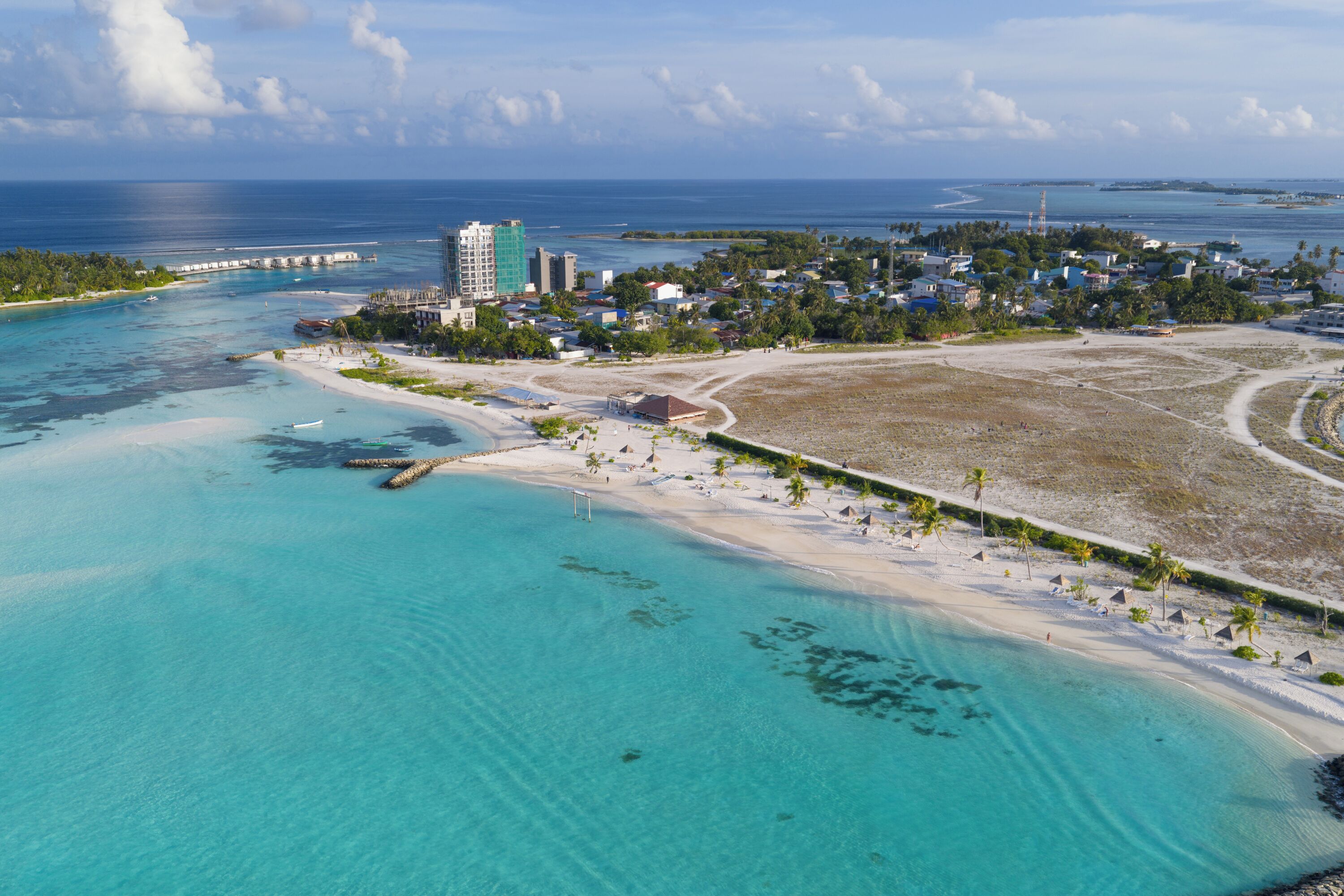 An aerial view of a coastal resort with a beach, clear turquoise waters, and a mix of buildings including a hotel and residential structures.