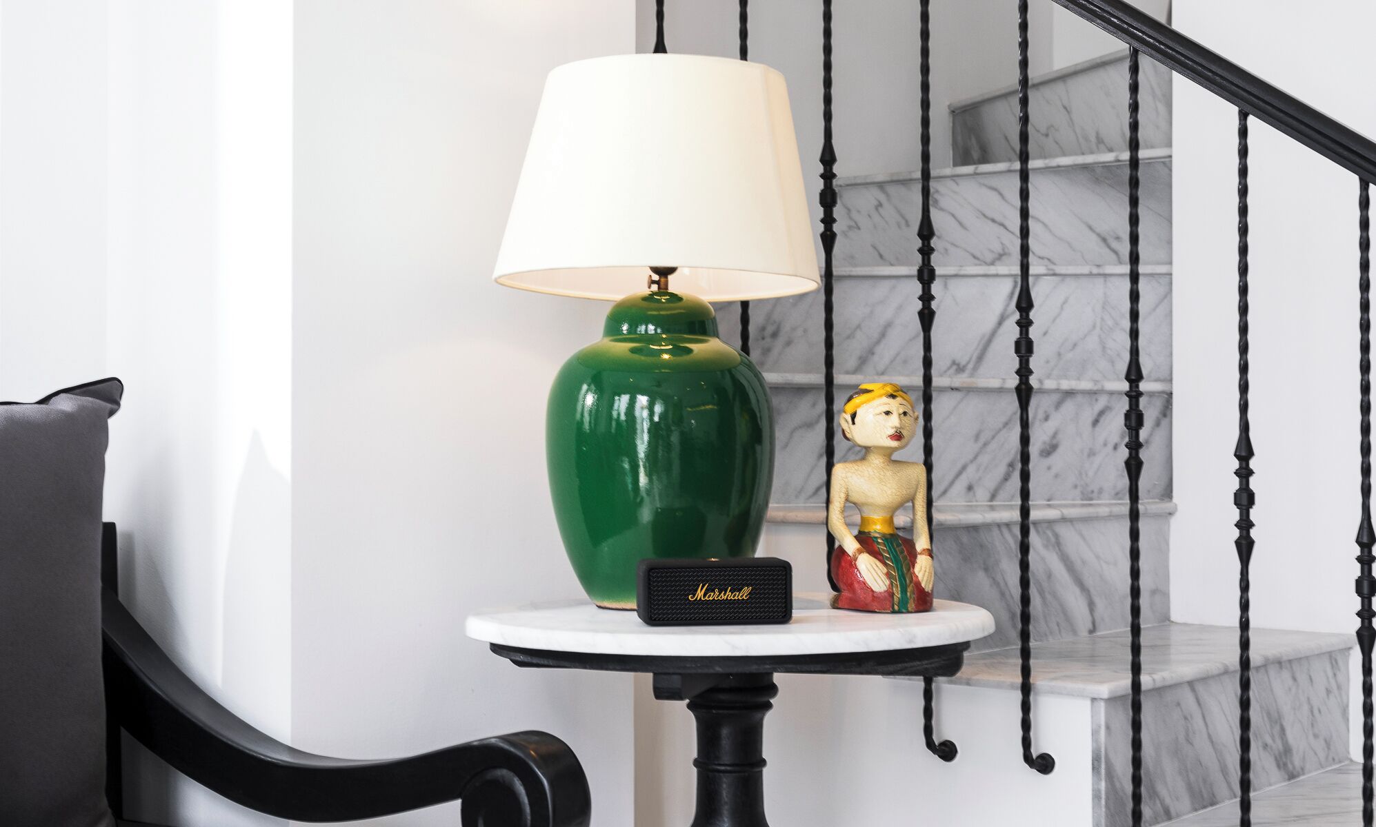 Eine grüne Lampe auf einem kleinen Tisch mit weißem Lampenschirm, eine kleine Figur und ein schwarzer Lautsprecher mit der Aufschrift 'Marshall' in einem Raum mit einer Marmortreppe.