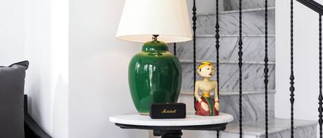 Eine grüne Lampe auf einem kleinen Tisch mit weißem Lampenschirm, eine kleine Figur und ein schwarzer Lautsprecher mit der Aufschrift 'Marshall' in einem Raum mit einer Marmortreppe.