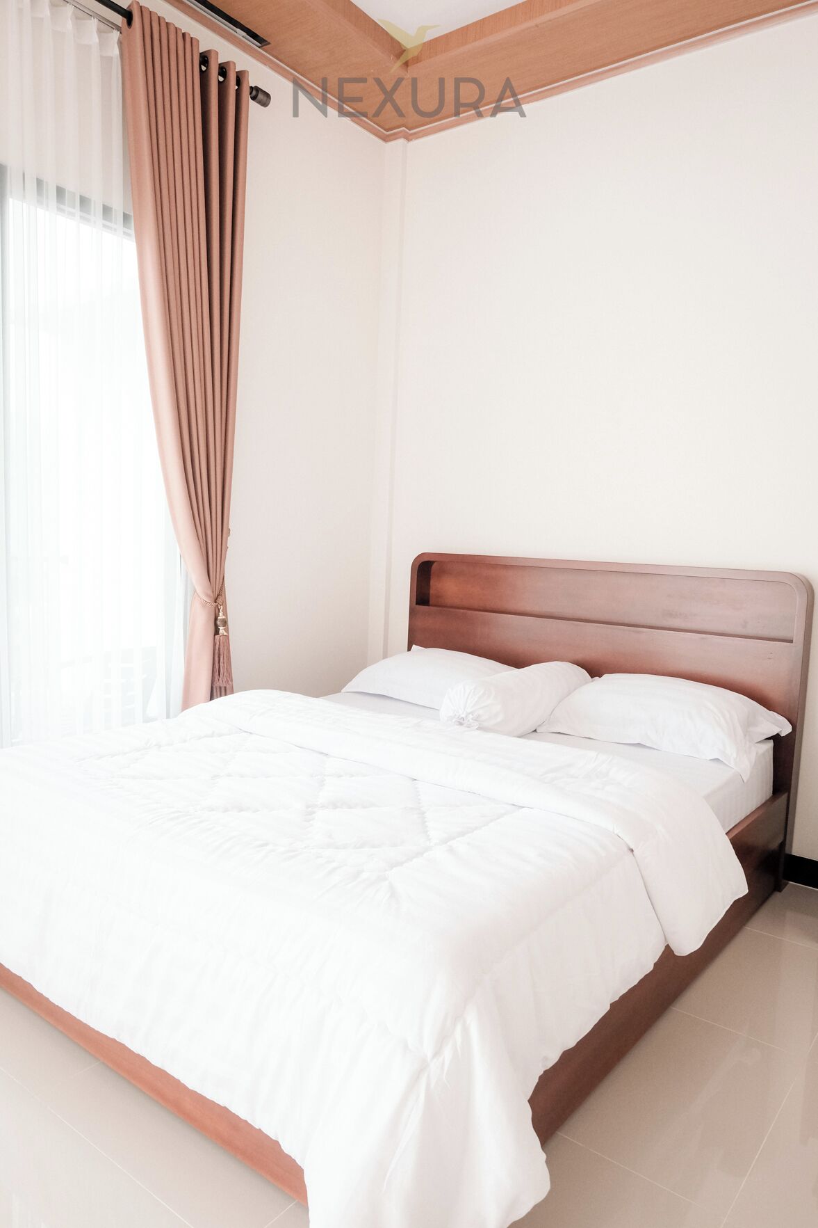 Un lit bien fait, avec du linge de lit blanc, dans une chambre dotée d’une tête de lit en bois et d’une fenêtre ornée de rideaux légers.