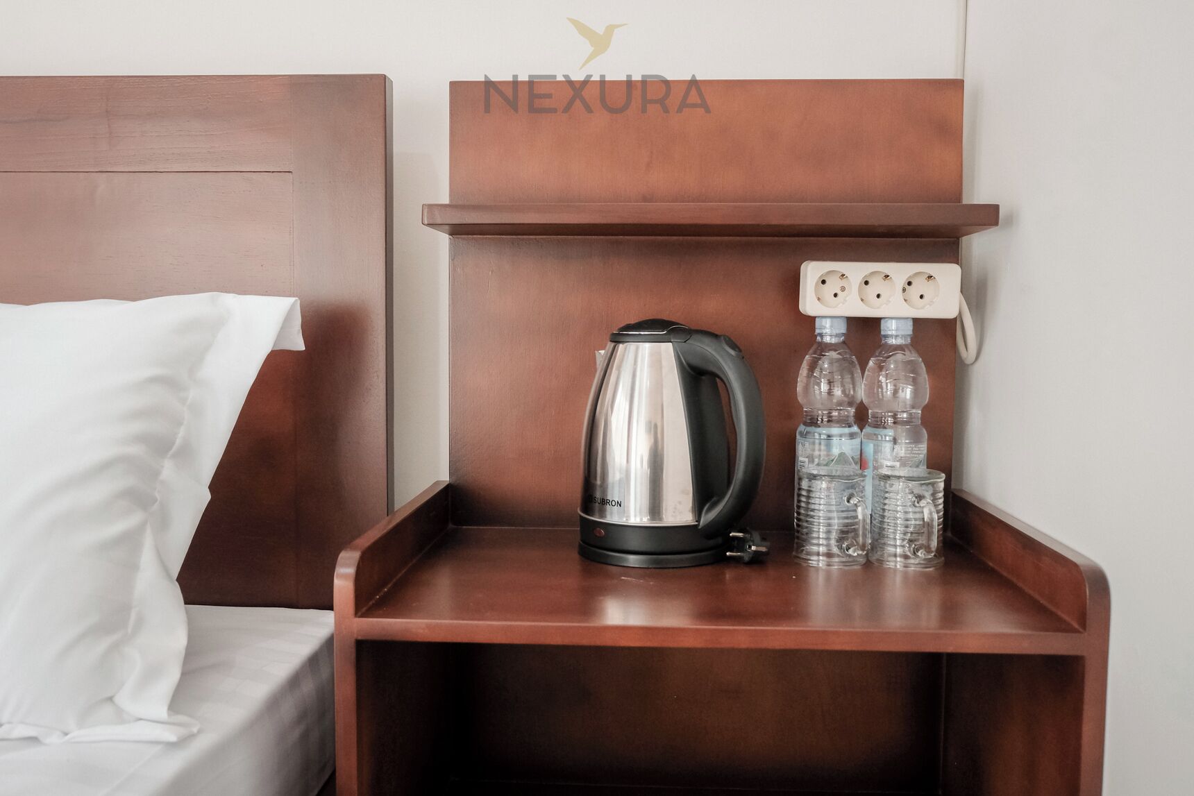 A wooden bedside table with a kettle, two water bottles, and a power outlet.