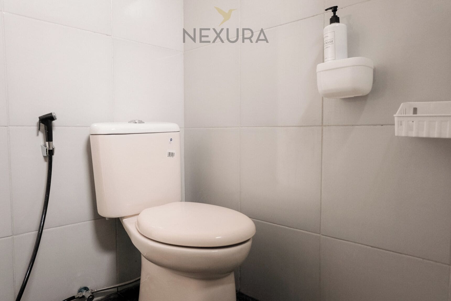 A white toilet with a black hose, a toilet paper holder, and a wall-mounted soap dispenser in a bathroom with white tiles.
