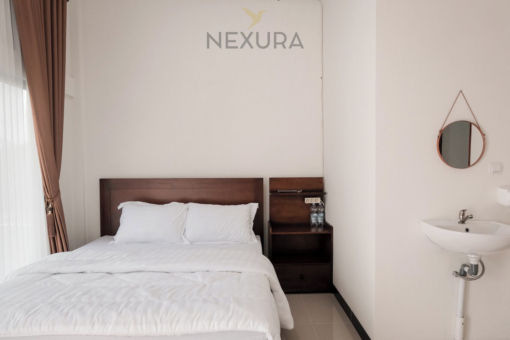 A hotel room with a bed, a nightstand with a mirror, and a wall-mounted sink.