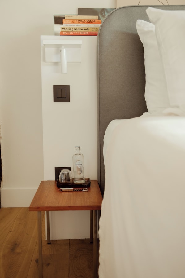 A neatly arranged hotel room with a bed, a bedside table with a bottle and glasses, a small nightstand with a lamp, and a stack of books.