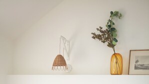 Een netjes opgemaakt bed met witte lakens en kussens, een klein houten nachtkastje, een wandlamp en een vaas met een plant.