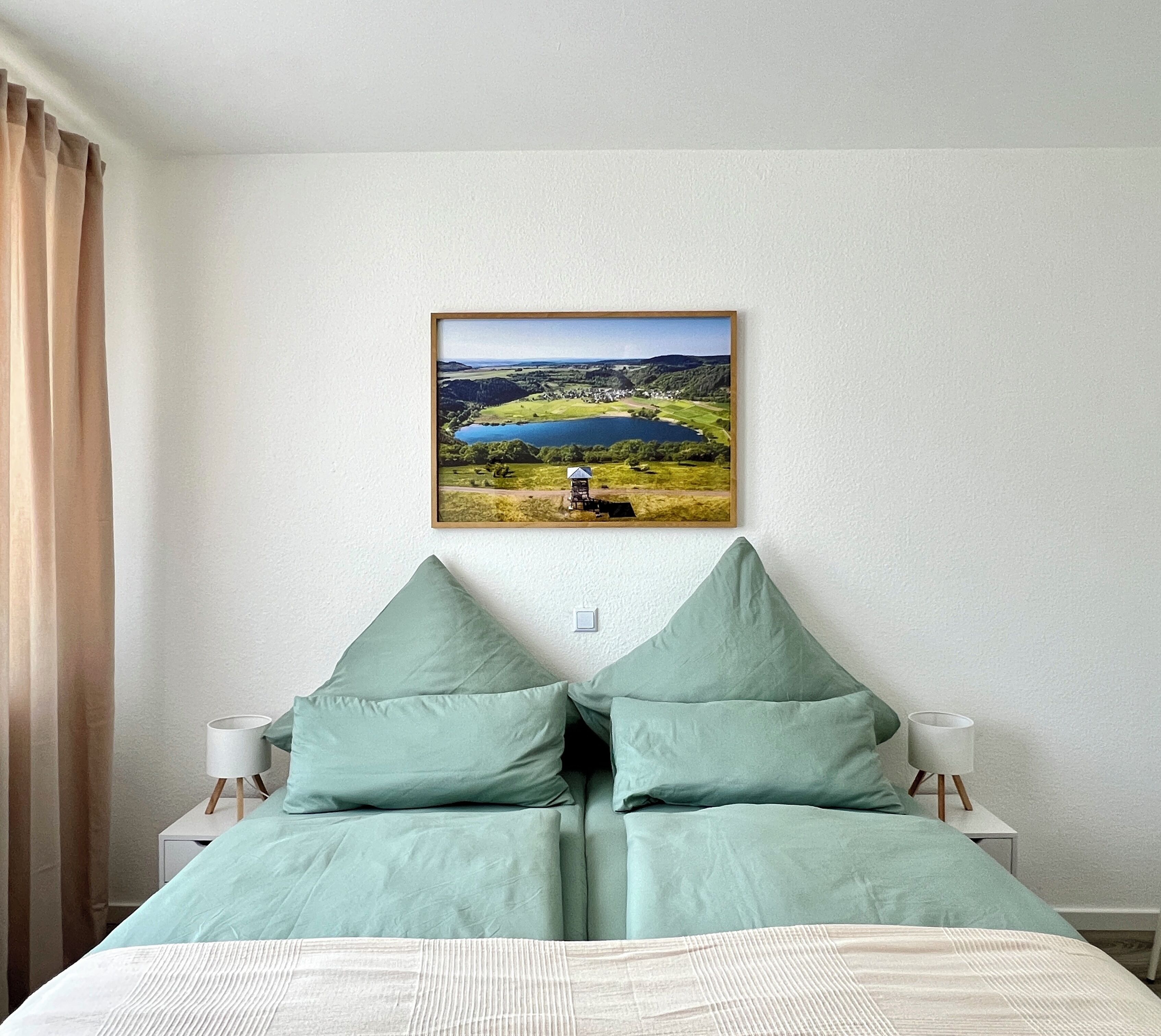 Ein Schlafzimmer mit einem Bett, grünen Kissen, einem gerahmten Landschaftsbild und Nachttischlampen.