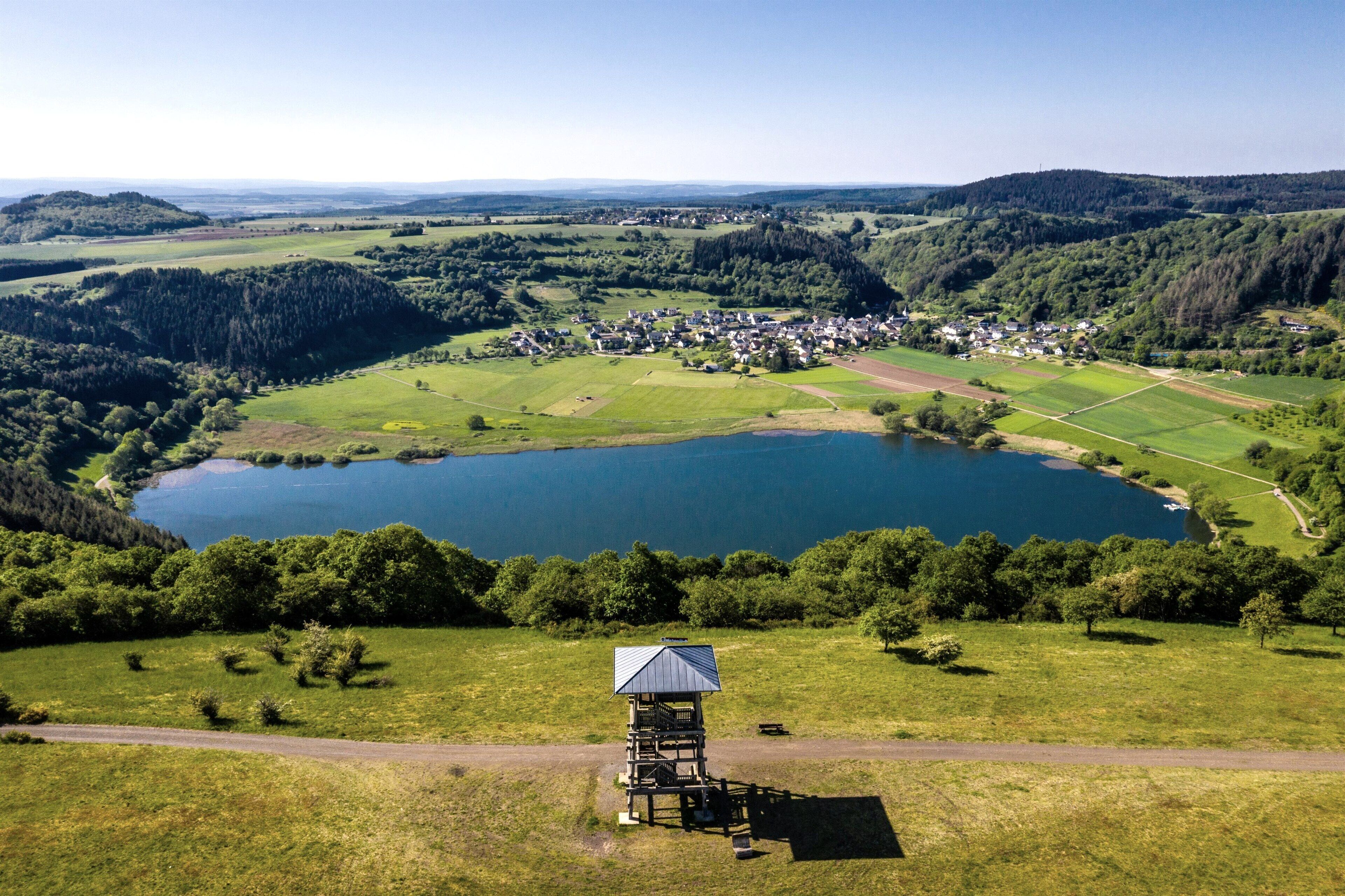 Blick auf ein Seeufer mit einem kleinen Holzbau am Wasser.