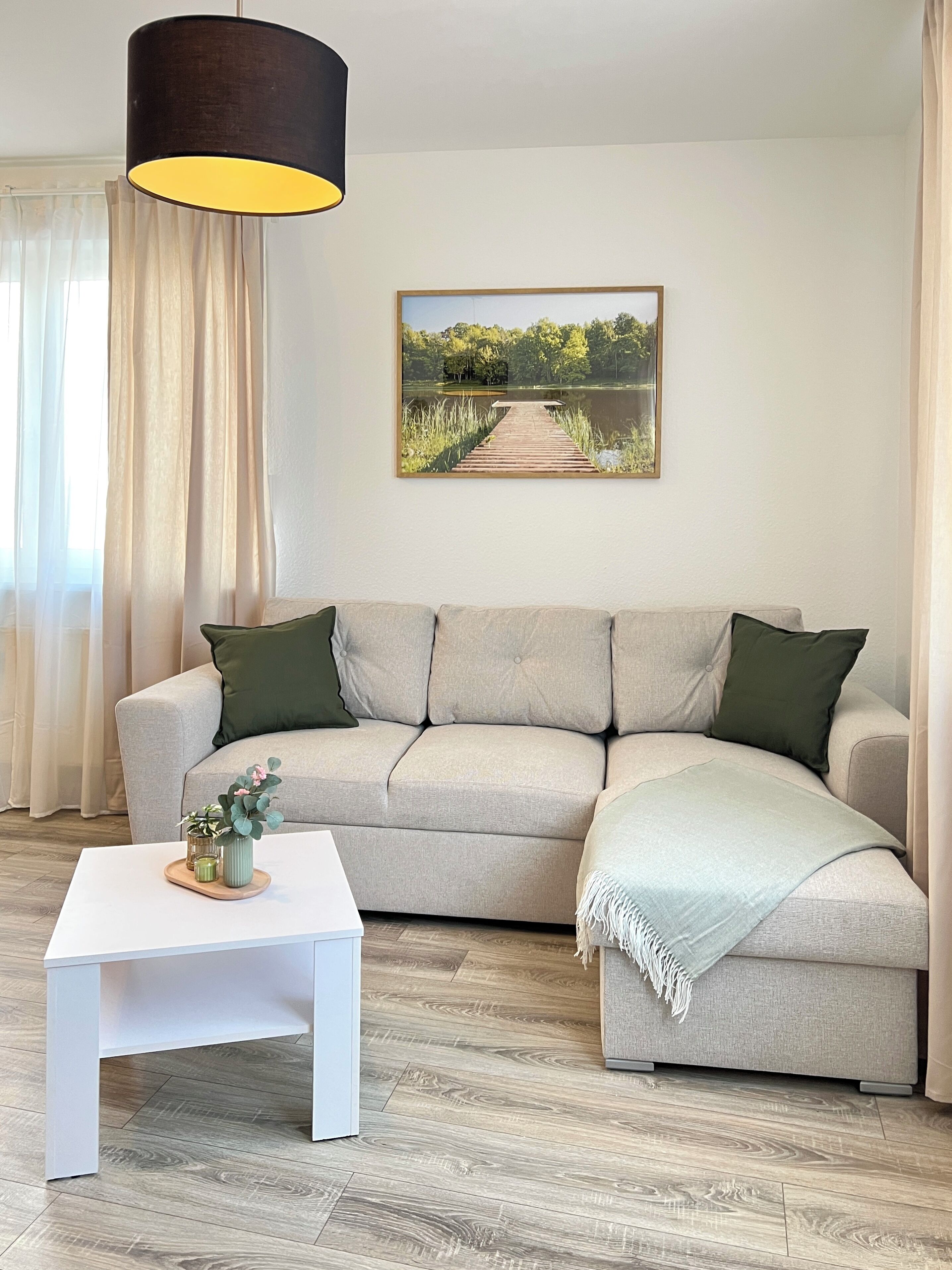 A living room with a beige sectional sofa, a white coffee table, and a painting of a lake and dock.