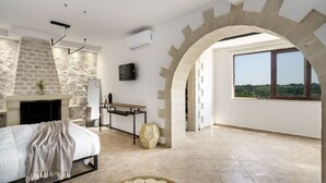 A bedroom with a stone fireplace, a bed with a white bedspread, a woven basket, and a large window with a view.