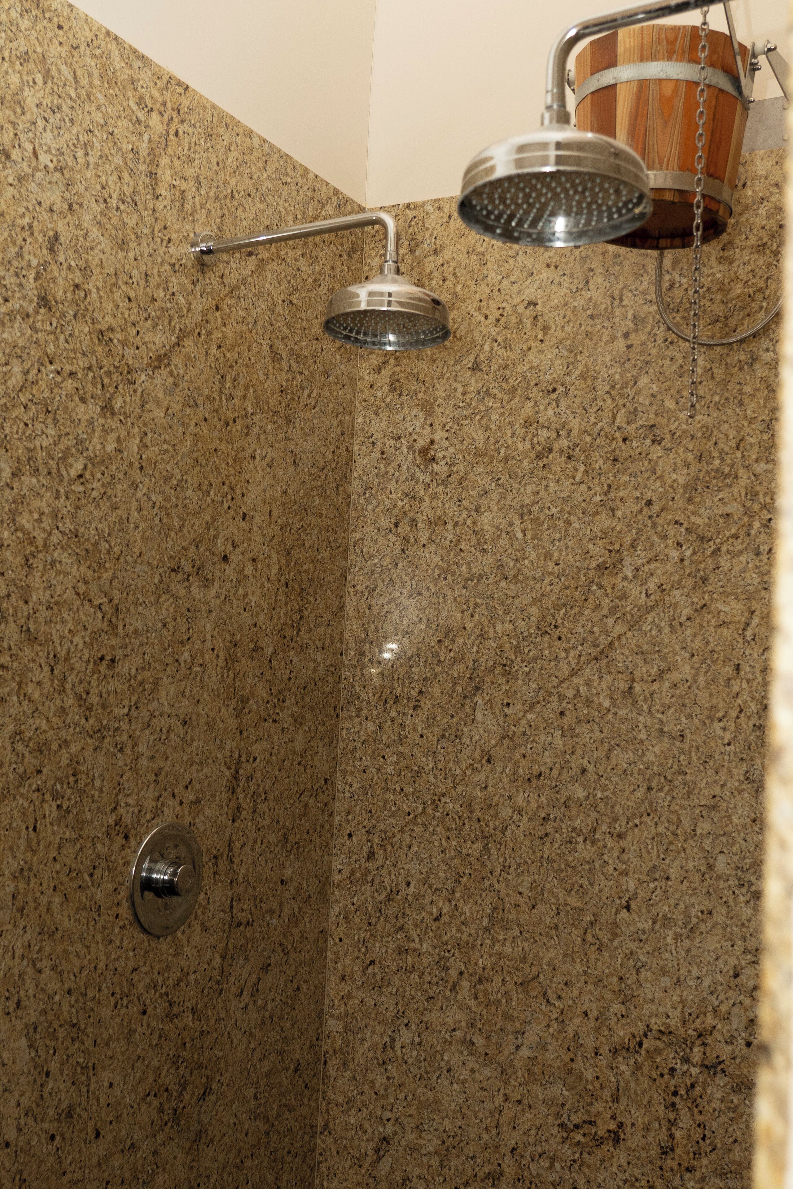 Un área de ducha con pared de granito, un cabezal de ducha metálico y un cubo de madera.
