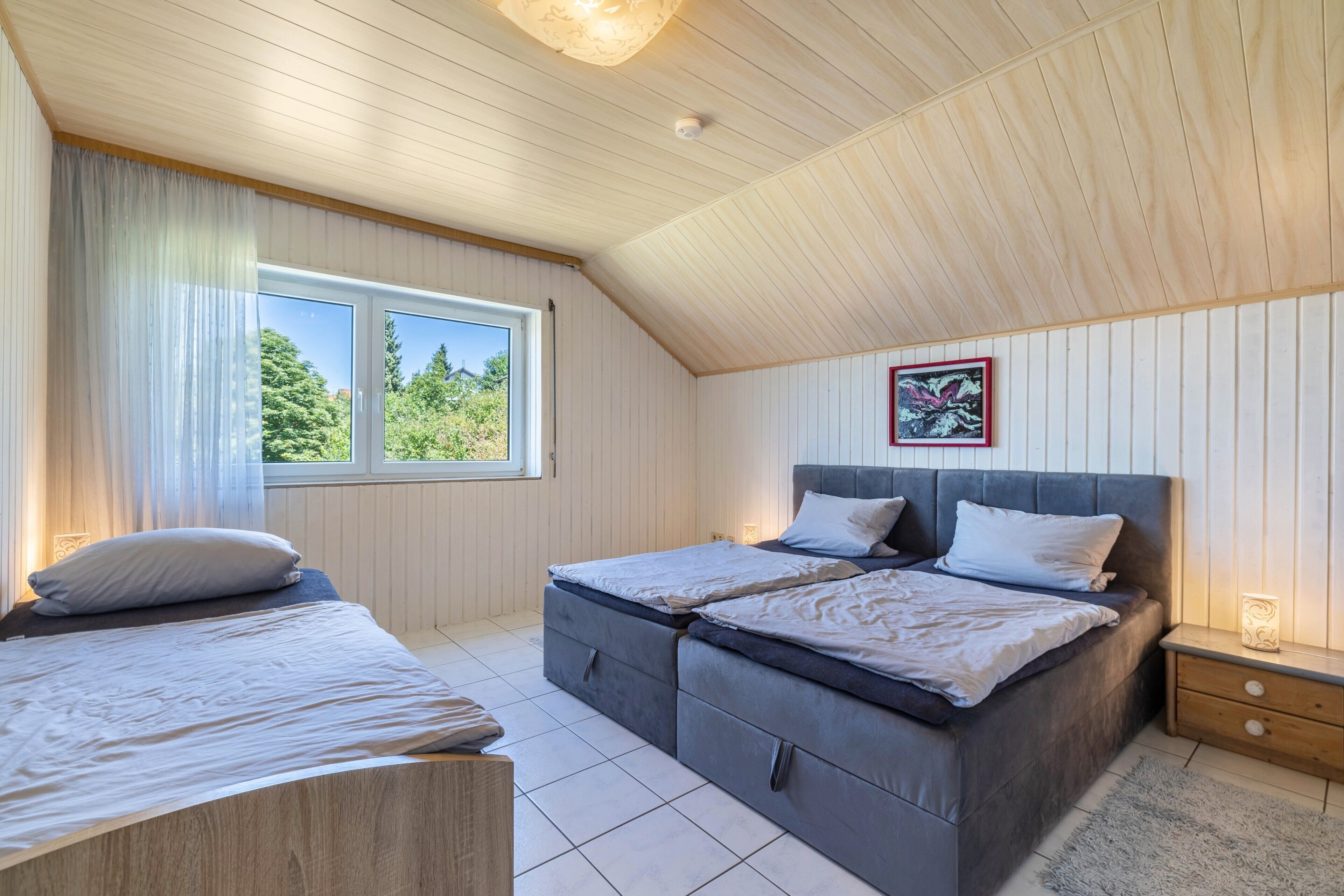 Un dormitorio con dos camas, una ventana con cortinas, una mesita de noche con lámpara y un cuadro en la pared.