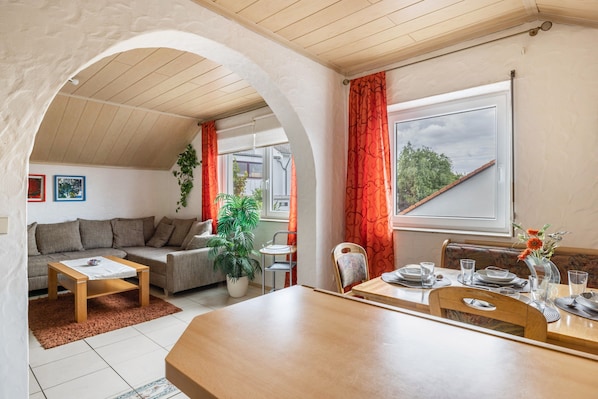Un espacio de sala acogedor con sofá, mesa de centro y mesa de comedor para dos, con vistas al exterior a través de un arco.