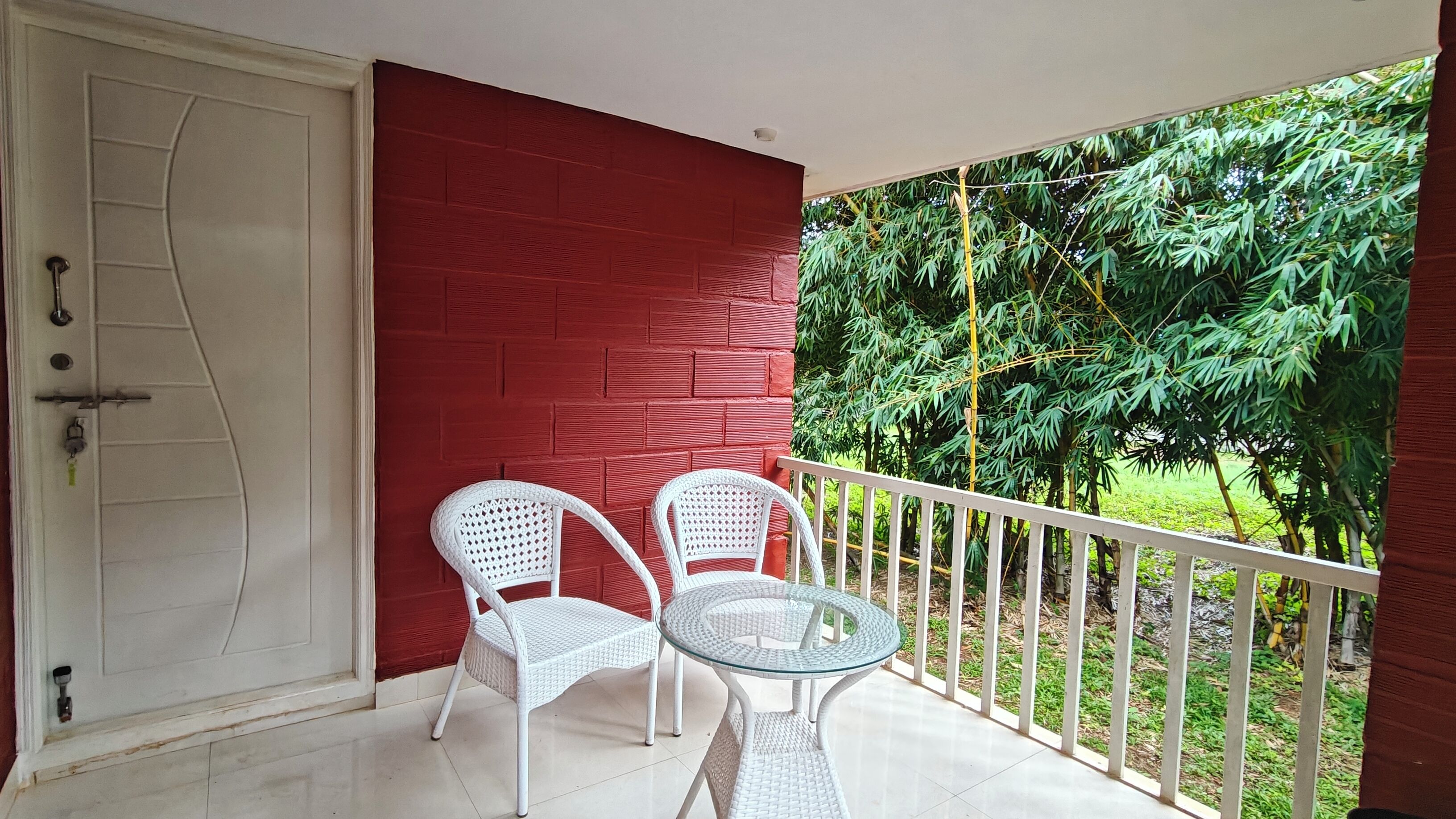 A balcony with two white wicker chairs and a glass table, a red wall, and a view of green foliage.