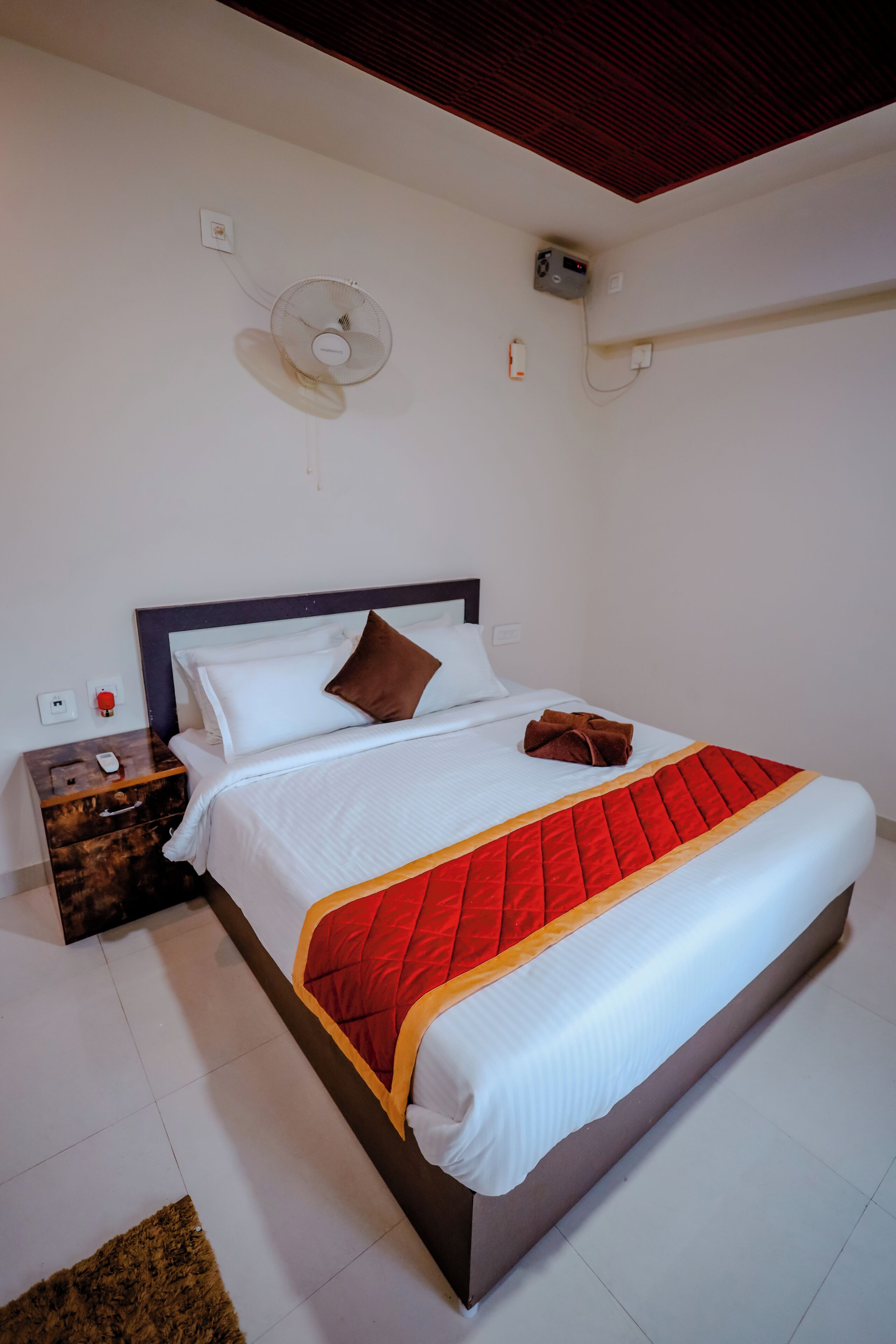 Una cama bien hecha con colcha roja y blanca, una mesita de noche de madera con un control remoto y un ventilador de pared.