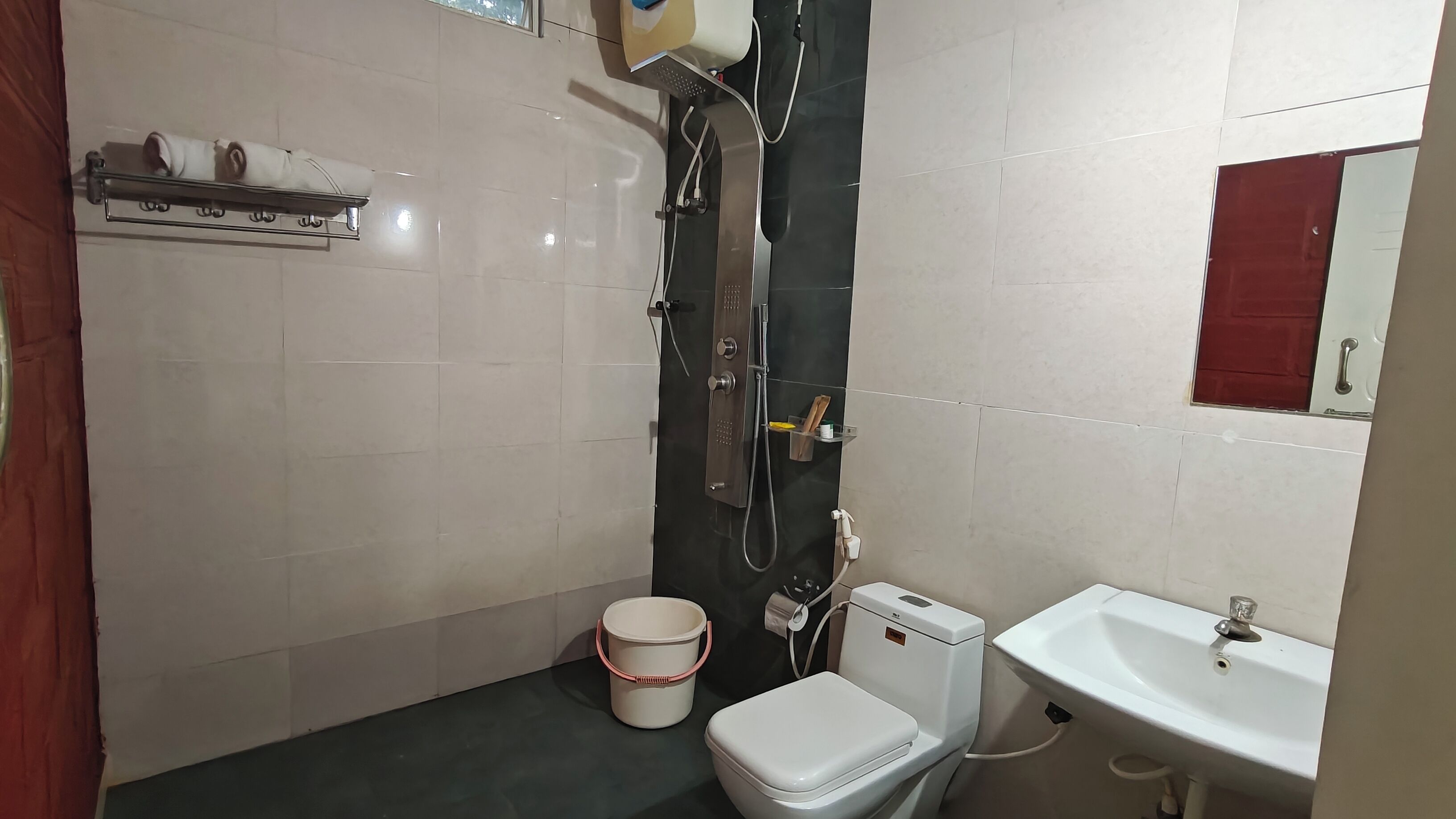 Un baño pequeño con inodoro, lavabo y ducha. Hay un toallero sobre el inodoro y una estantería con artículos de aseo.