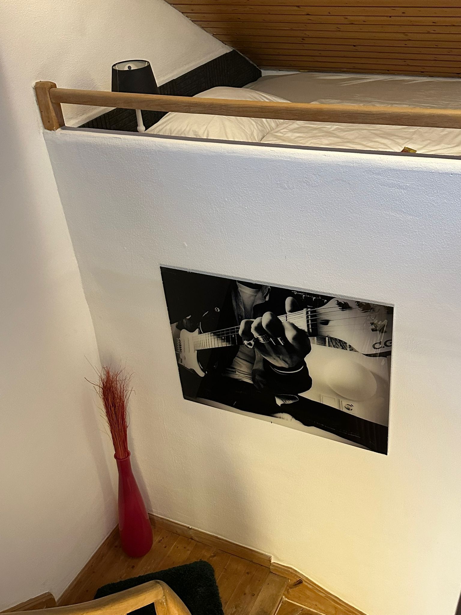 Un dormitorio con una cama litera, una fotografía en blanco y negro enmarcada de una persona tocando la guitarra, un florero rojo y piso de madera.
