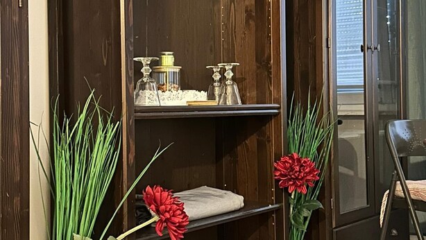 Eine dunkle Holzkommode mit Regalen, einige mit dekorativen Gegenständen und Kristallgläsern. Auf dem Boden neben der Kommode befinden sich zwei Vasen mit Grünpflanzen und roten Blumen.