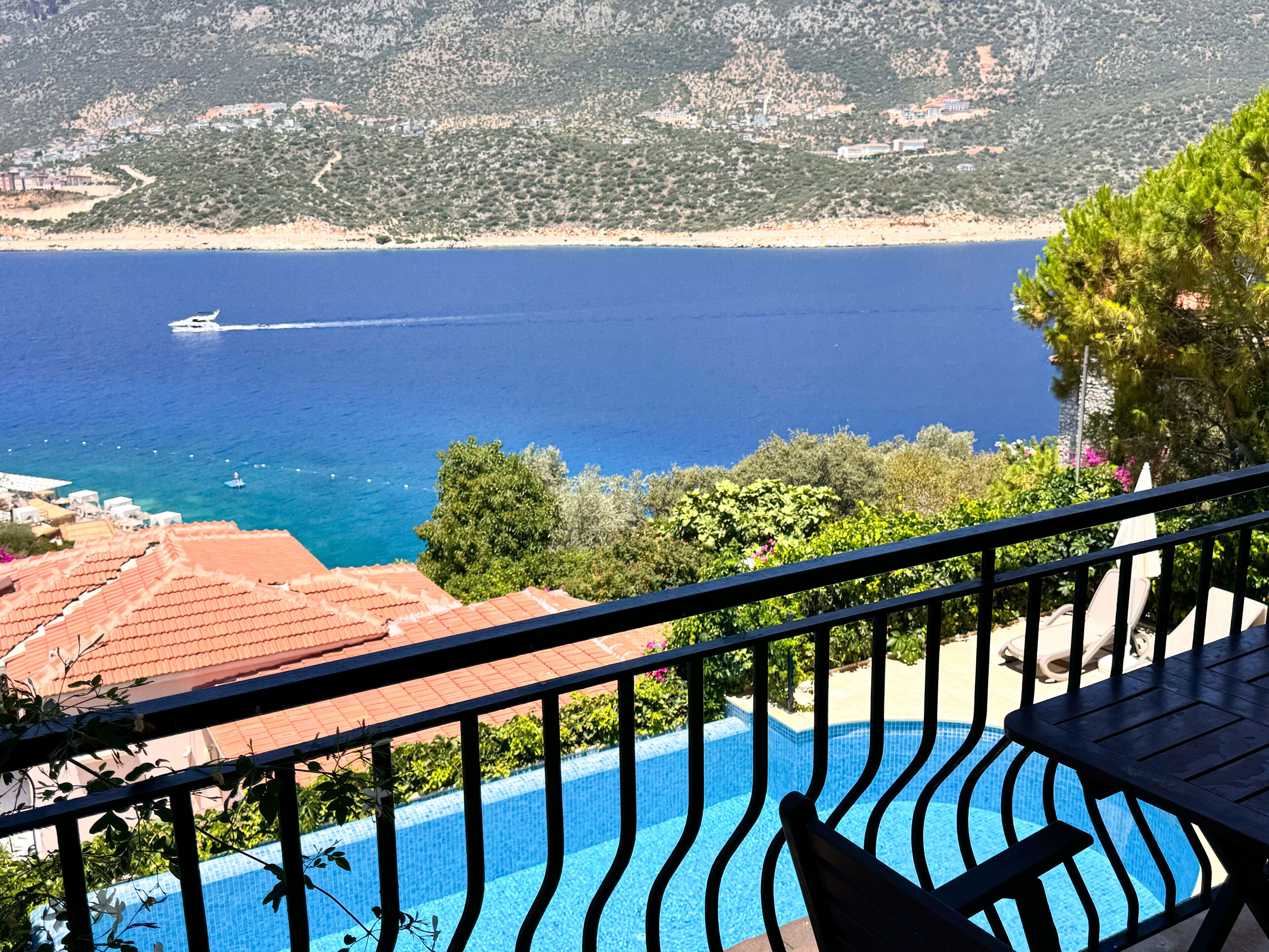 A balcony with a view of a swimming pool, a boat on the sea, and a coastal landscape.