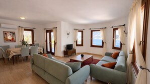 A living room with a sofa, TV, dining table, and windows with curtains. - Villa Noa Amazing Sea view (Kas)